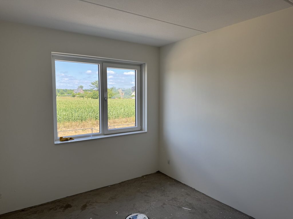 Lege slaapkamer in een nieuwbouwhuis met gladde witte wanden en een raam dat uitkijkt op een maisveld